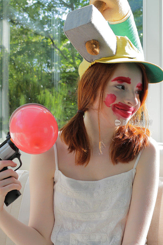 14 / 15岁的红发少女坐在阳光明媚的温室里玩气球爆破游戏，玩气球爆破游戏，戴着小丑脸和傻帽